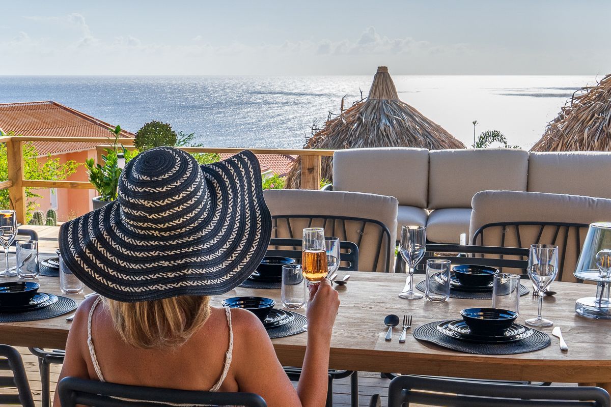 Een vrouw met een hoed heft haar glas, zittend aan een gedekte tafel met uitzicht op de oceaan.