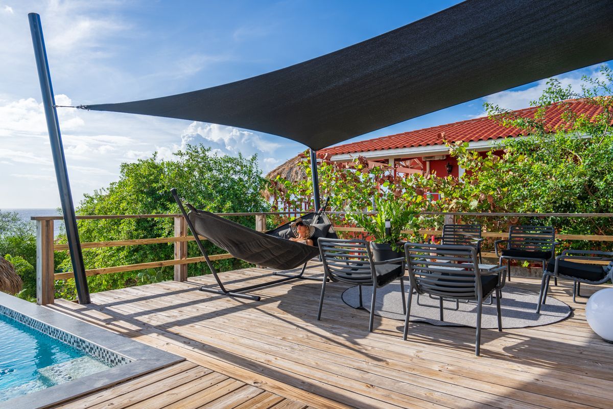 Terras met hangmat, tafel, stoelen en schaduwdoek bij een zwembad; groen en een gebouw op de achtergrond.