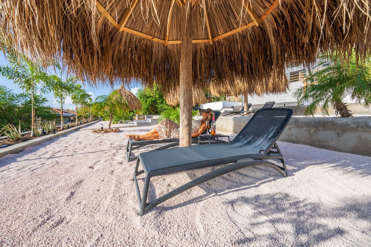 Ligstoelen onder een rieten parasol op een grindterras. Mensen ontspannen en zonnebaden.