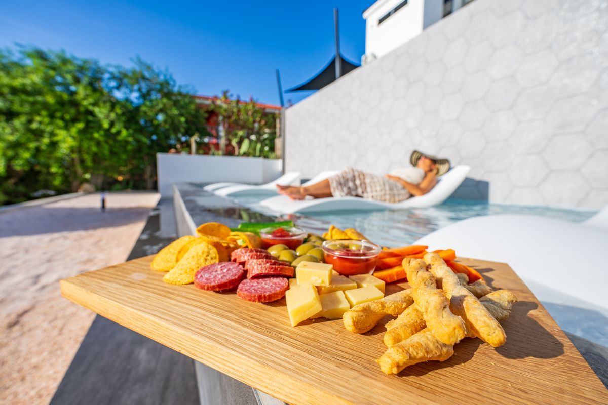 Een plank met charcuterie en hapjes bij een zwembad, met iemand die aan het relaxen is.