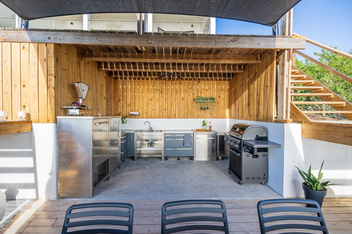 Buitenkeuken met roestvrijstalen apparatuur, grill en houten wanden. Zonnescherm boven het hoofd.