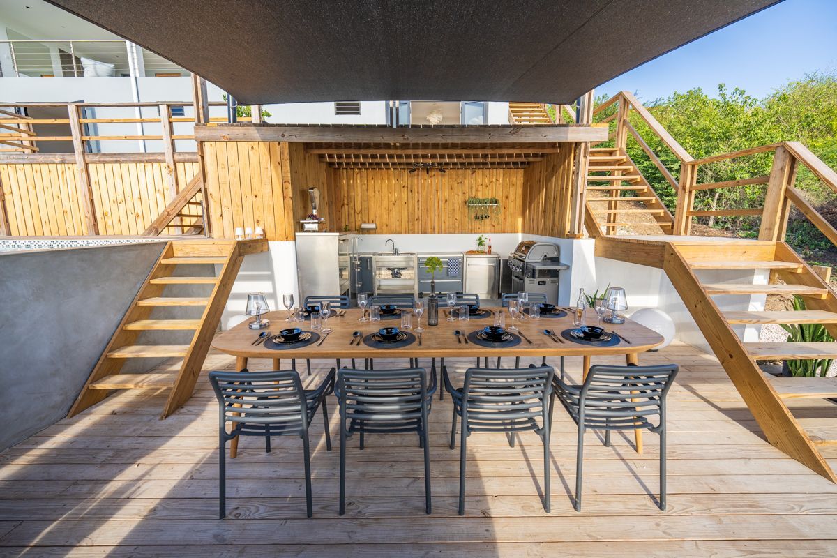 Eetgedeelte buiten met een lange tafel gedekt voor een maaltijd; houten terras, trap en keuken op de achtergrond.