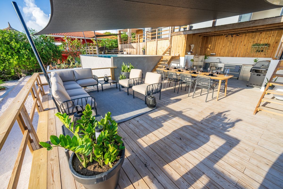 Buitenterras met zitgedeelte, eettafel en ingebouwde buitenkeuken. Zwart zonnescherm erboven, zwembad op de achtergrond.