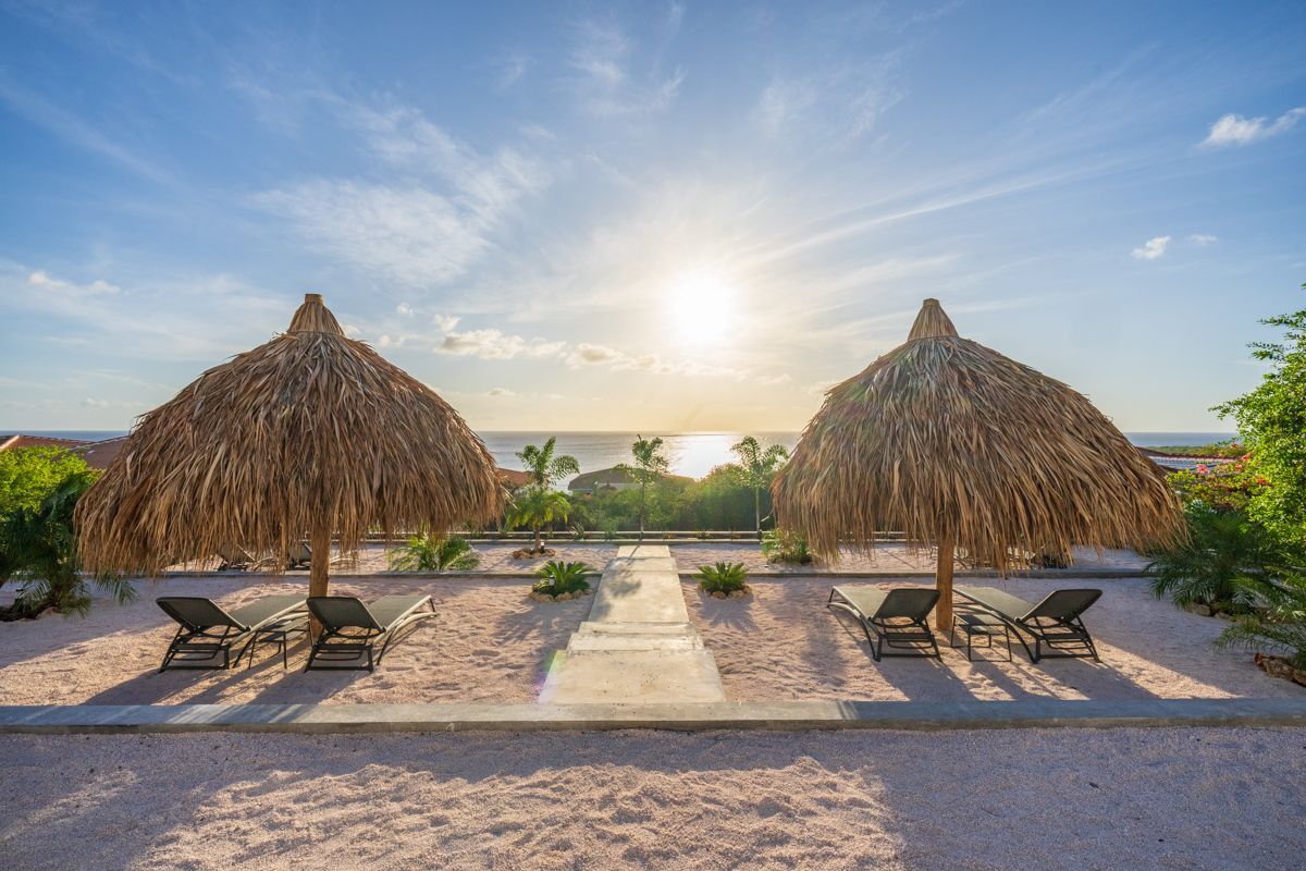Twee rieten parasols bieden schaduw aan ligstoelen op een strand, met uitzicht op de oceaan onder een stralende zon.