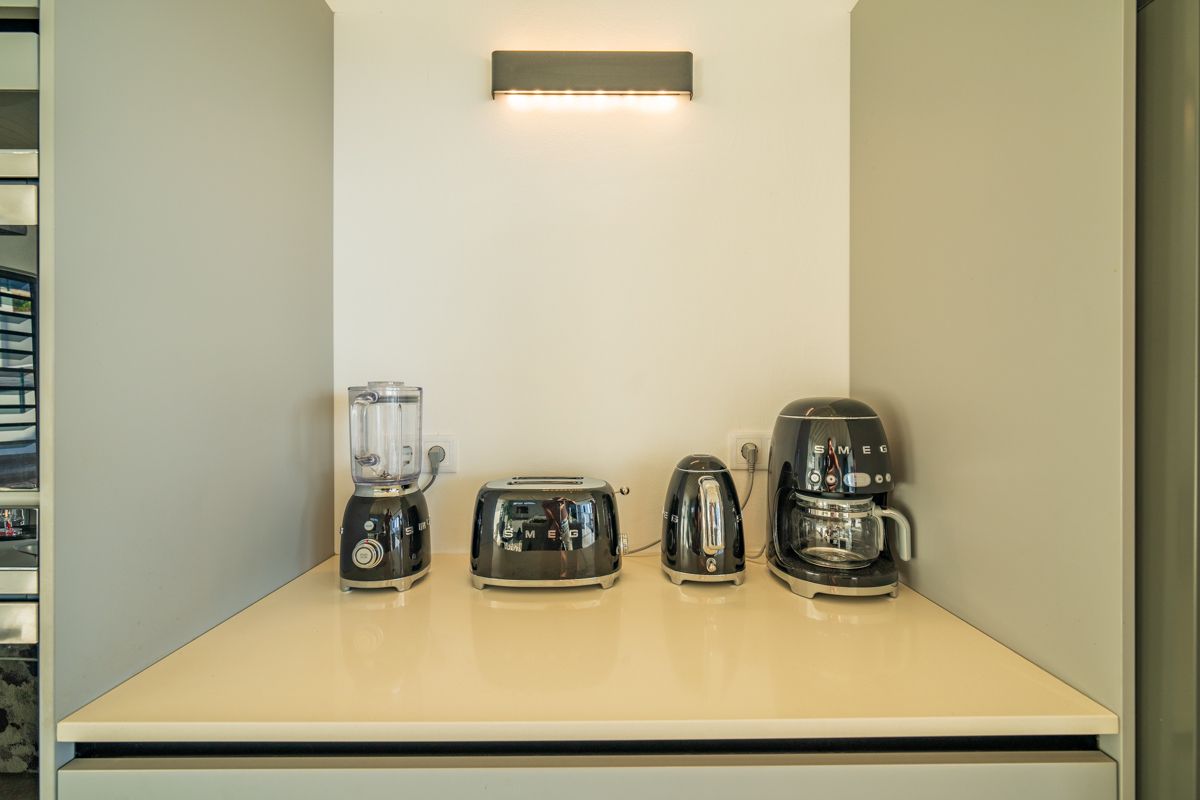Ingebouwd keukenblad met blender, broodrooster, waterkoker en koffiezetapparaat. Grijs en wit, onder een wandlamp.