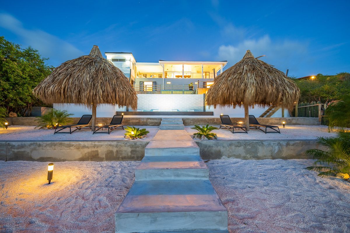 Villa aan het strand met rieten parasols, ligstoelen en een pad naar het gebouw, met een blauwe schemering in de lucht.