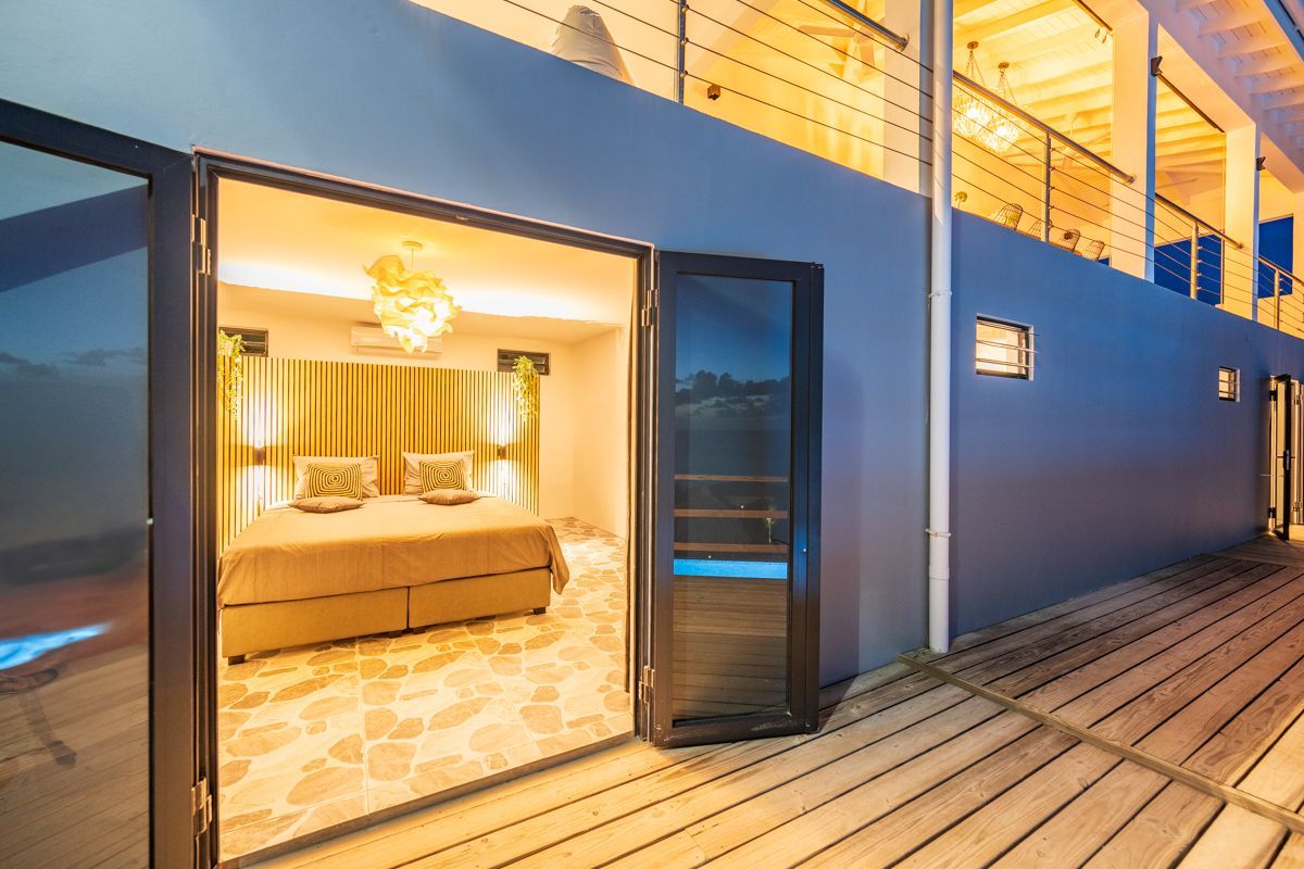 Slaapkamer met openslaande glazen deuren naar een terras. Lichte kamer, neutrale kleuren, bed en decoratieve plafondlamp.