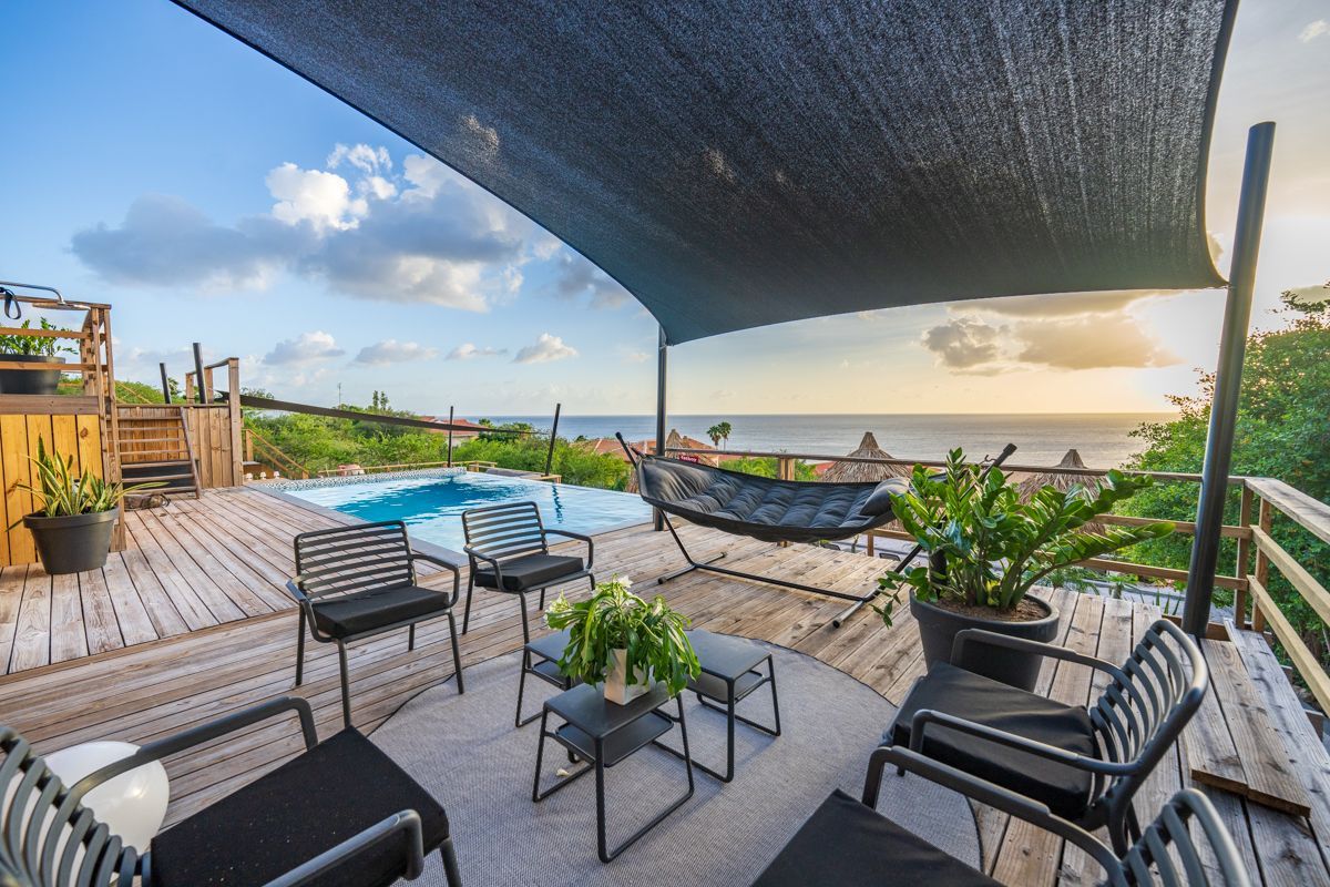 Houten terras met een zwembad, stoelen en een hangmat met uitzicht op de oceaan, onder een parasol.