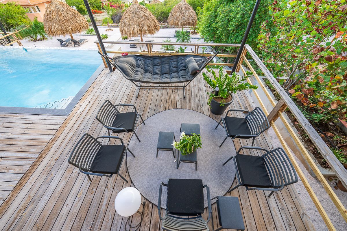 Terras met zwarte stoelen rond een kleine tafel, een hangmat en een zwembad. Houten vlonder in een zonnige buitenomgeving.