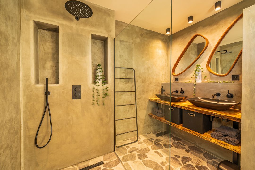 Moderne badkamer met betonnen muren, glazen douchecabine, zwarte kranen, houten wastafelmeubel en decoratieve spiegels.