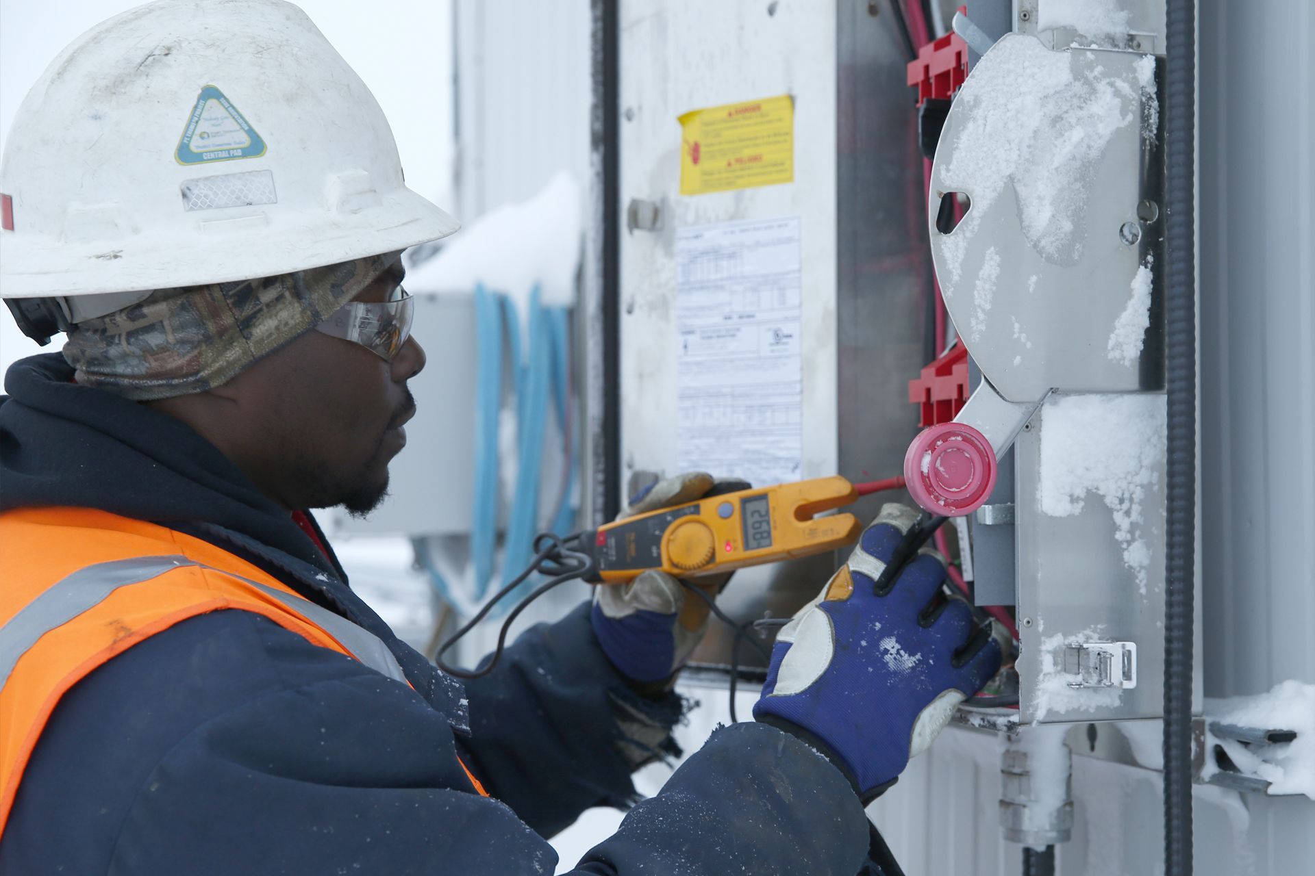 A man wearing a hard hat and safety vest is working on an electrical box in the snow.