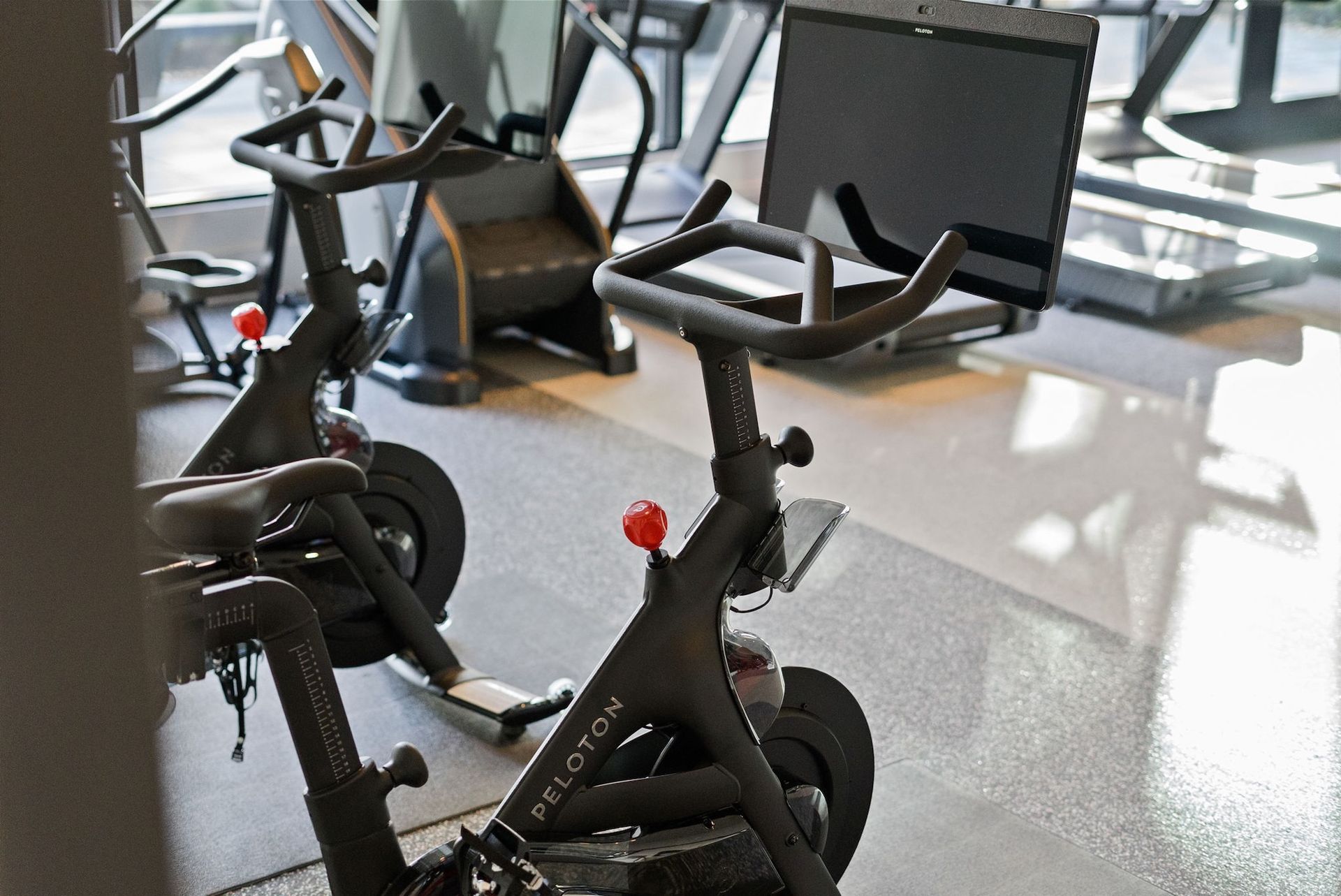 Two Peloton stationary bikes in a gym with sunlight reflecting off the floor.