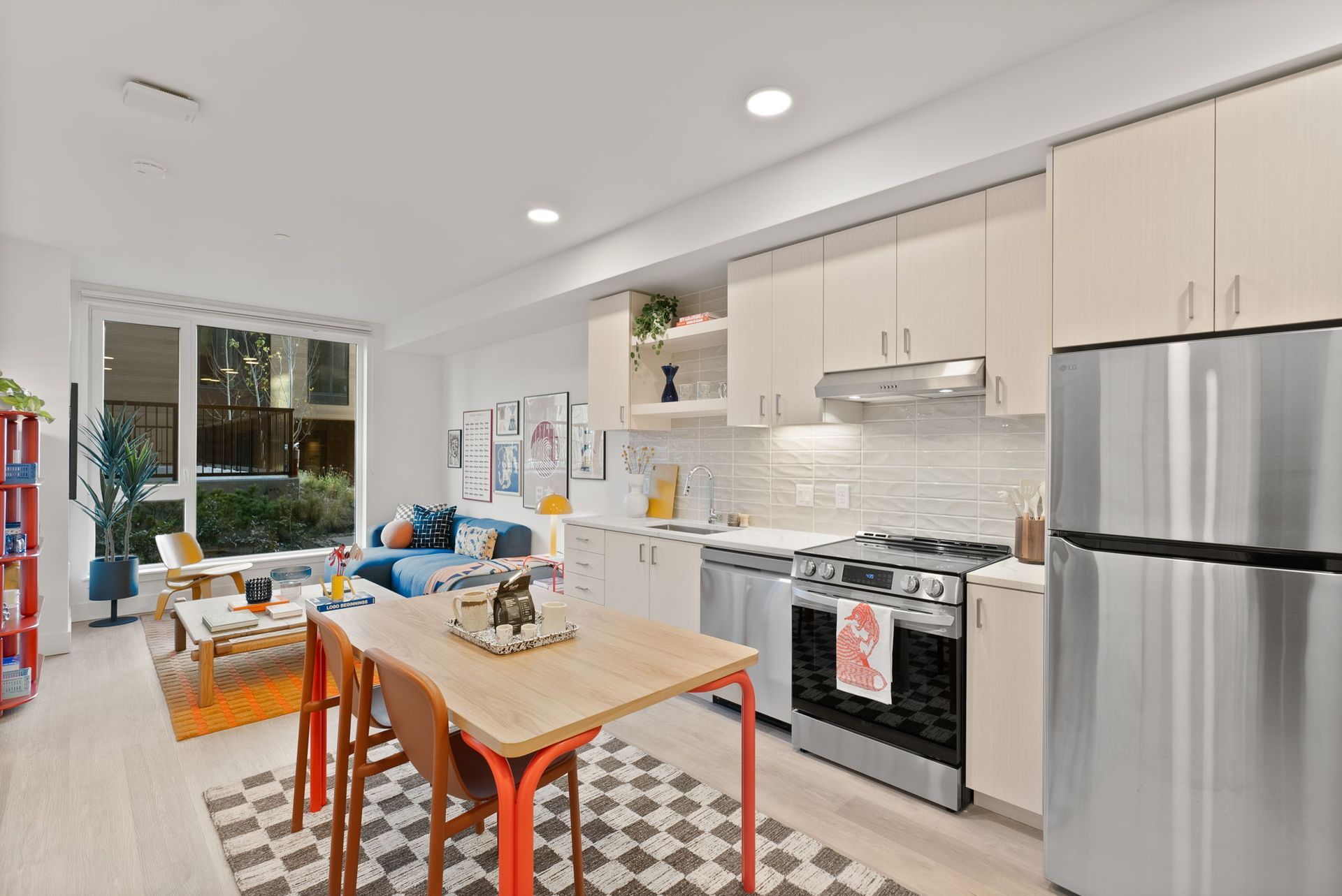 Open-concept kitchen and living area, light wood cabinets, orange accents, window overlooking greenery.