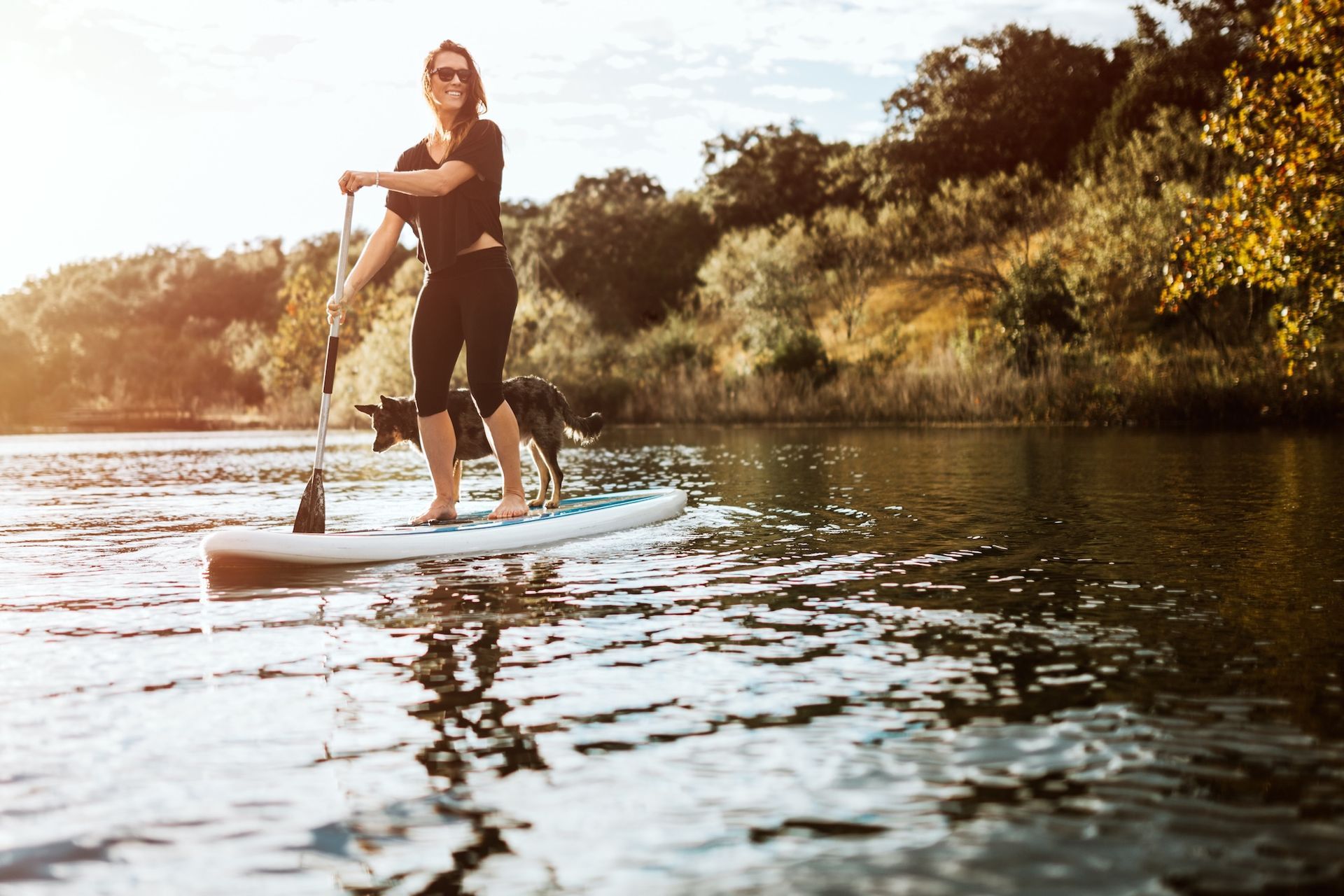 Woman paddleboarding on a lake with a dog. The setting is sunny and surrounded by trees, with rippling water.