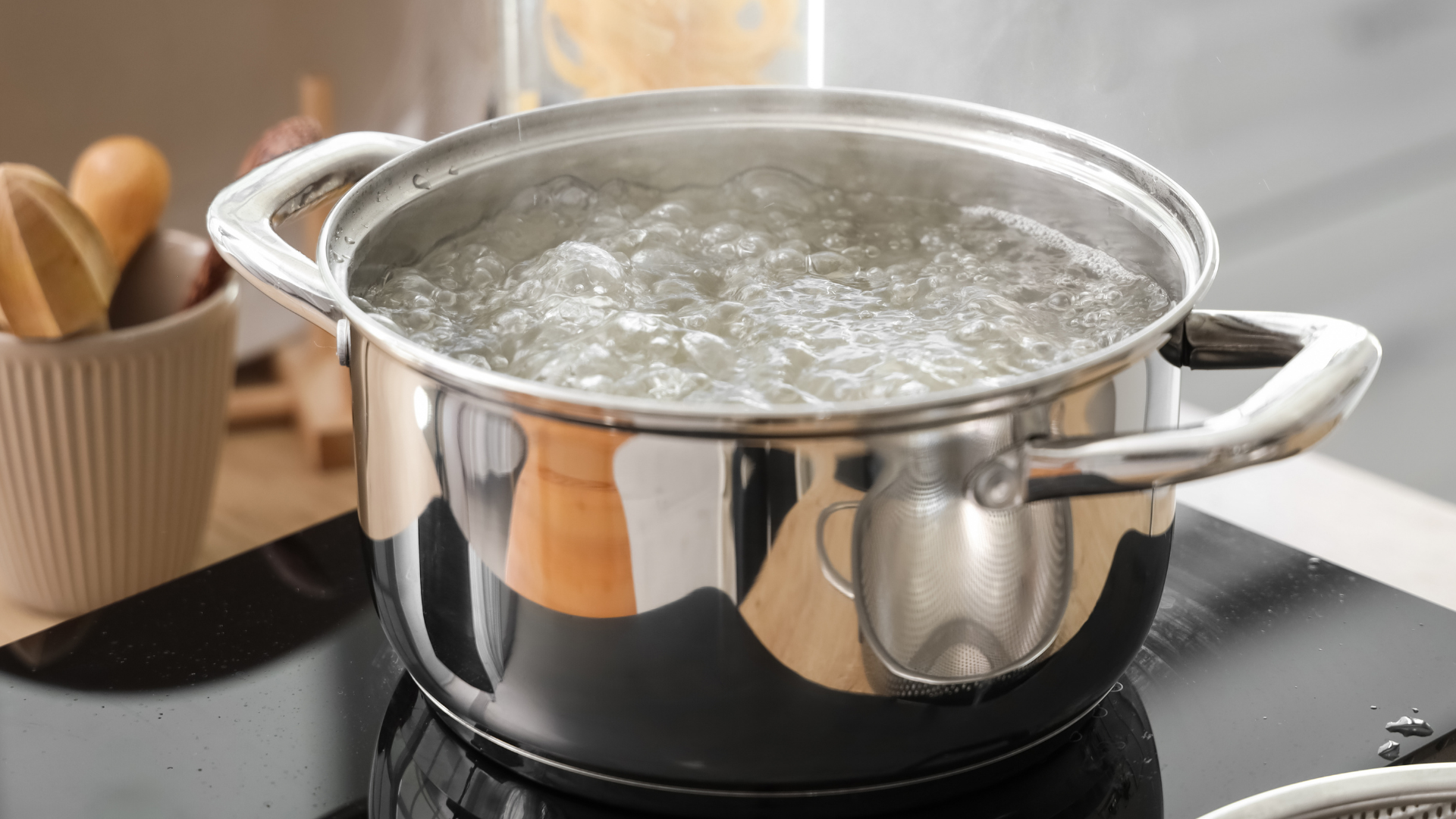Stainless steel pot of boiling water on a stovetop, with kitchen utensils beside it.