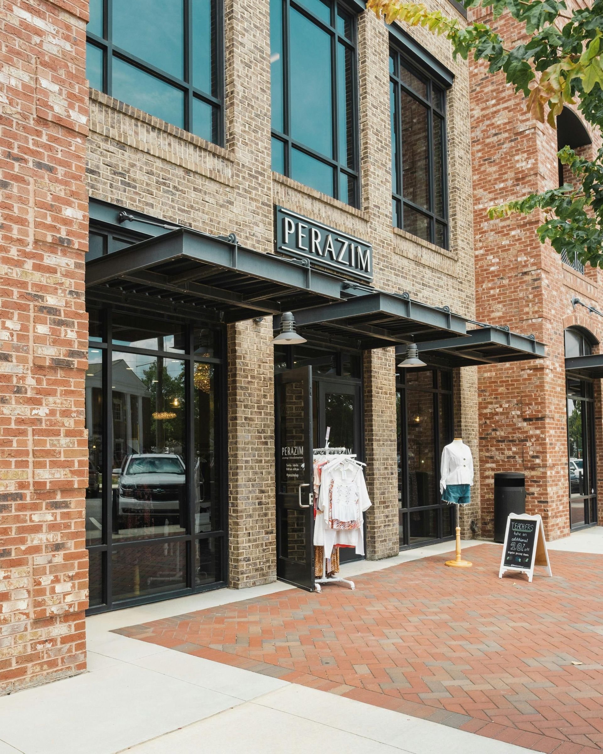 Brick storefront with large windows, clothing displays, and a sidewalk sign outside.