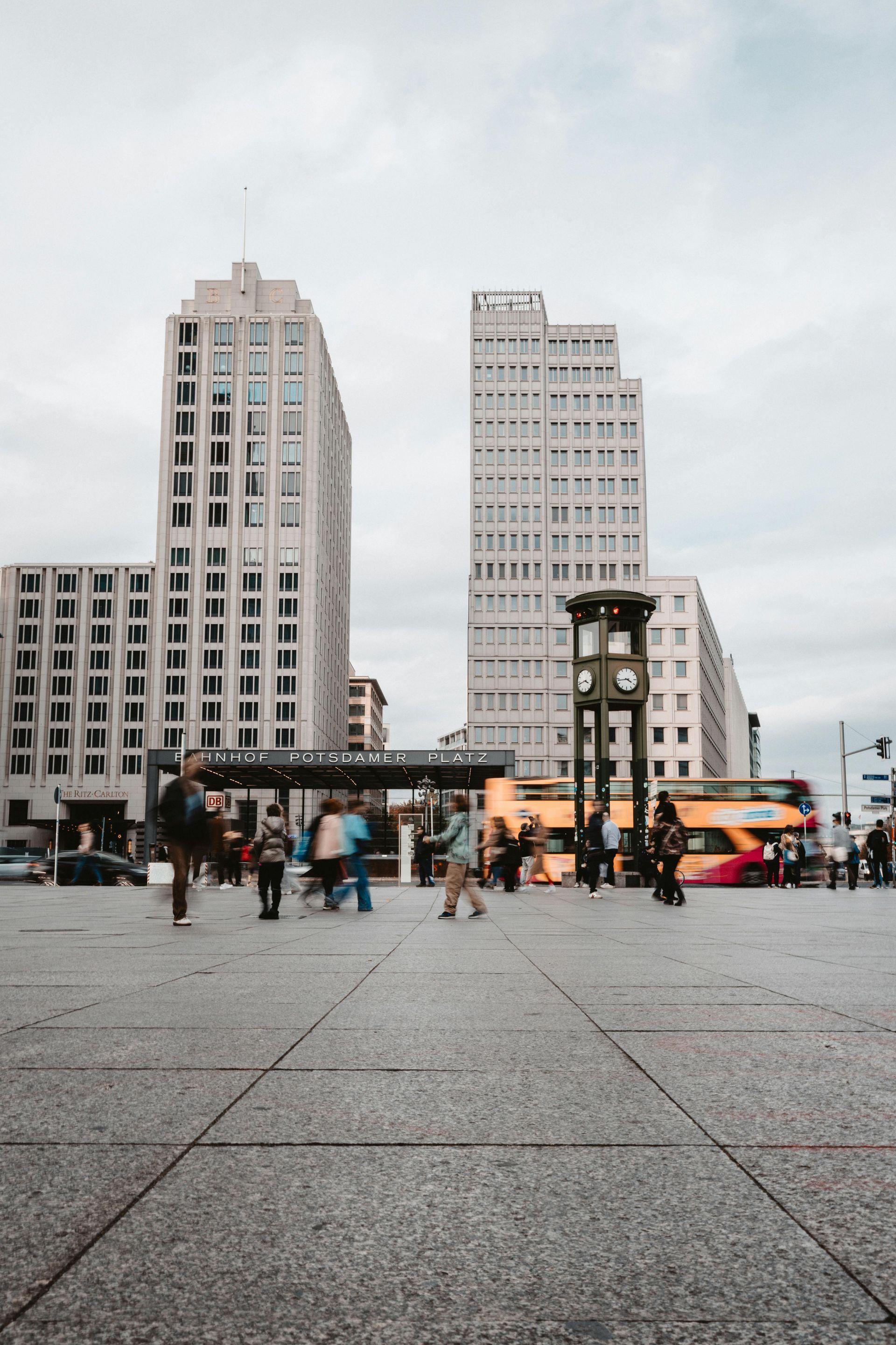 City square with tall buildings, pedestrians, and a red bus under an overcast sky
