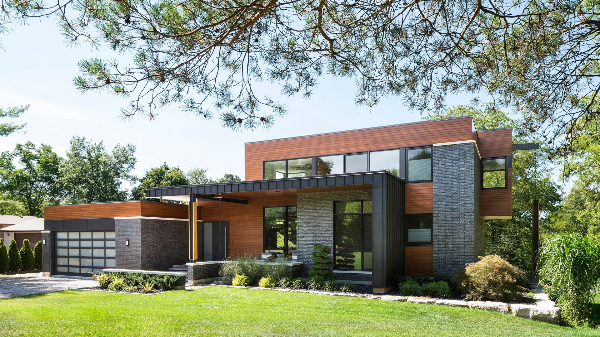 Modern house with wood and stone exterior, green lawn, and trees under a blue sky.