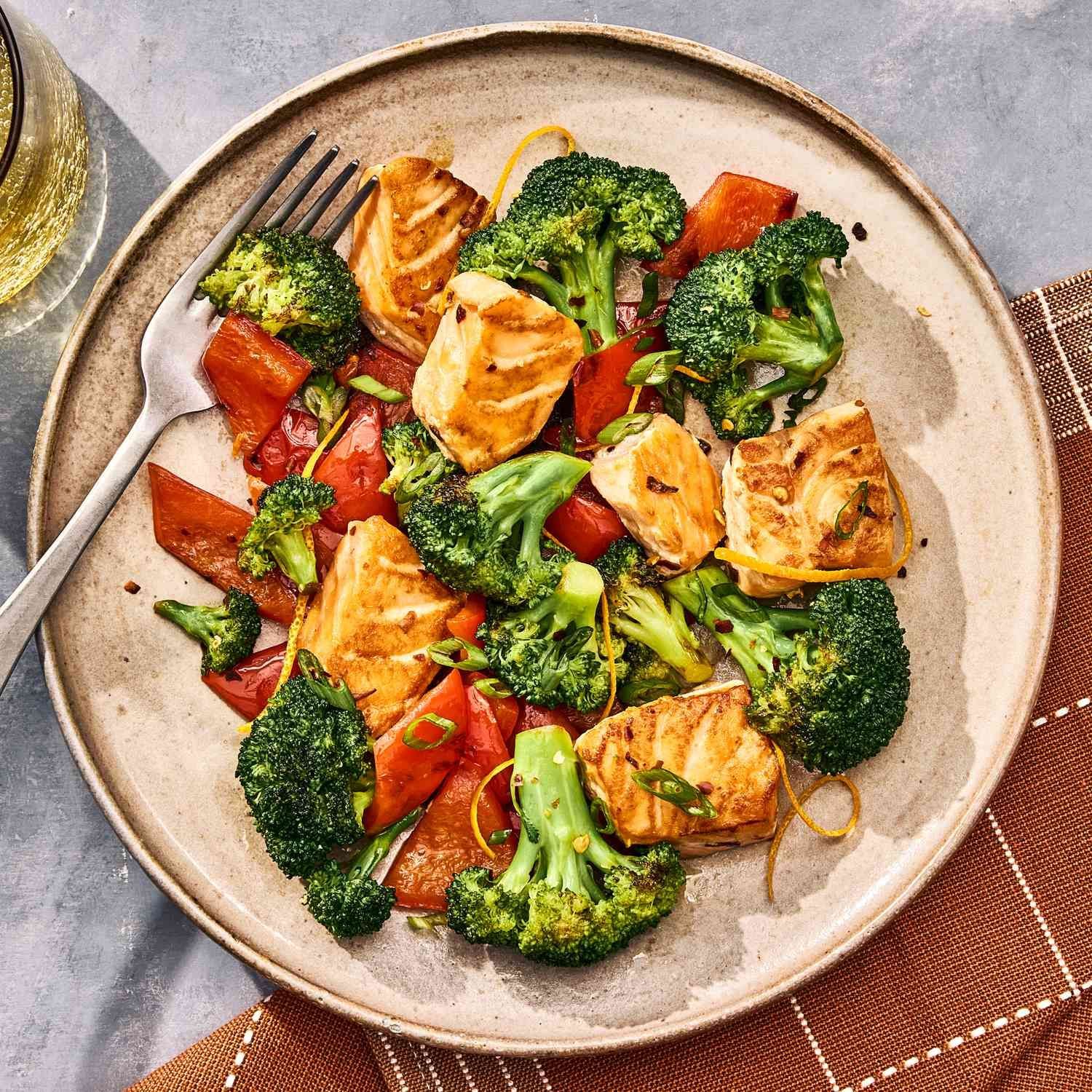 Een bord met zalm, broccoli en geroosterde paprika's op een tafelkleed.