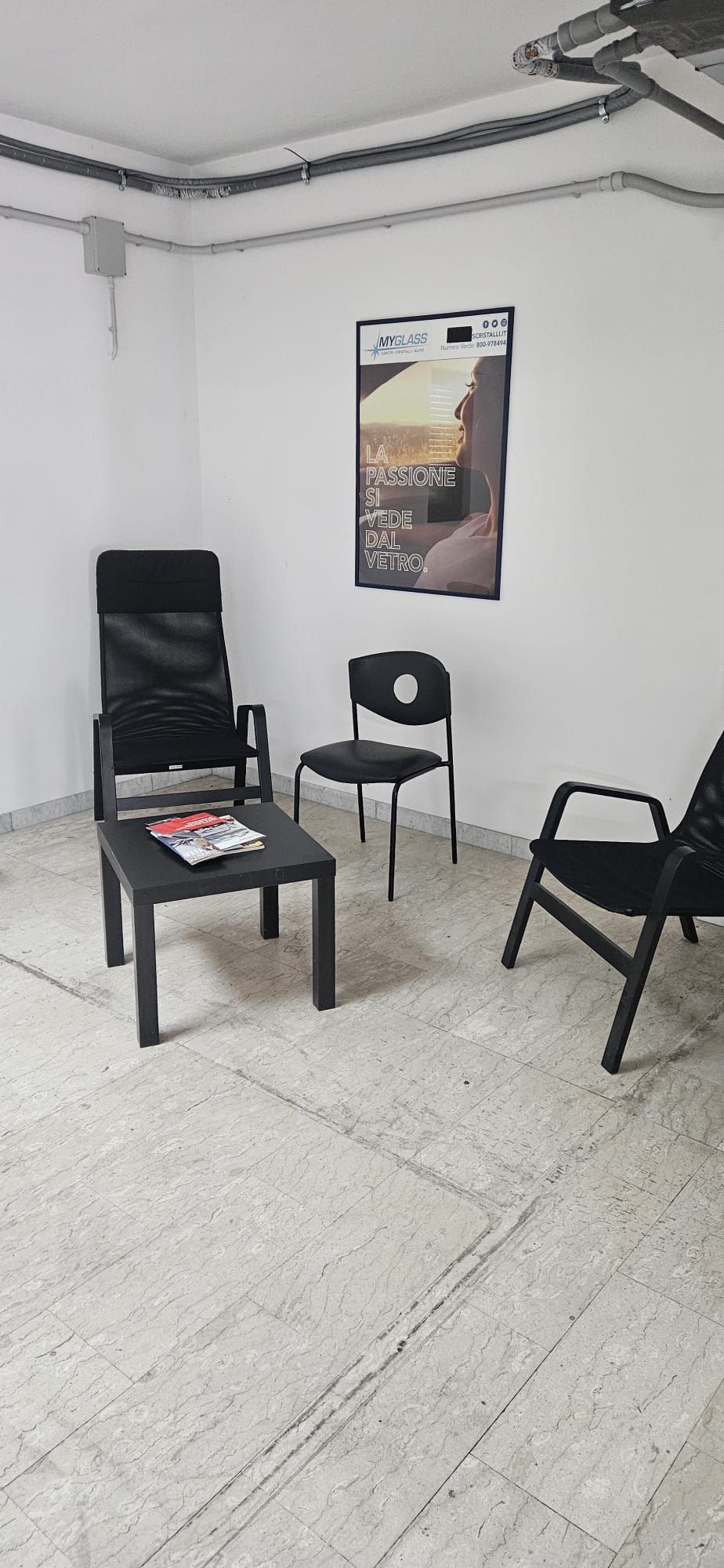 Una stanzetta spoglia con tre sedie nere, un tavolino basso e un poster appeso al muro.