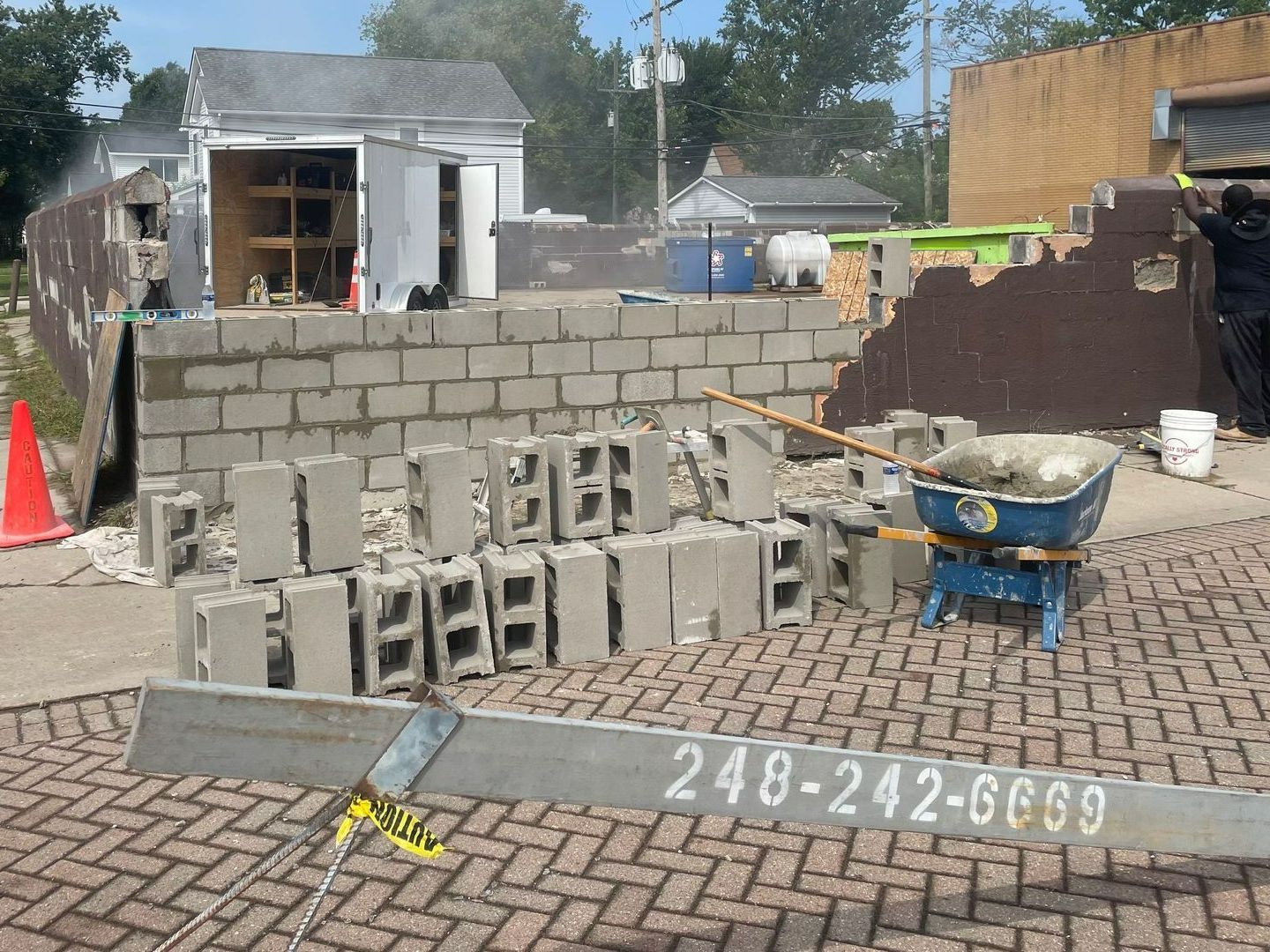 Construction site with cinder blocks, a wheelbarrow, orange cone, and partially built brick wall.