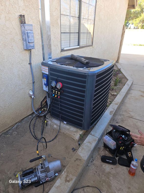 A technician services an outdoor air conditioning unit connected to a vacuum pump with manifold gauges.