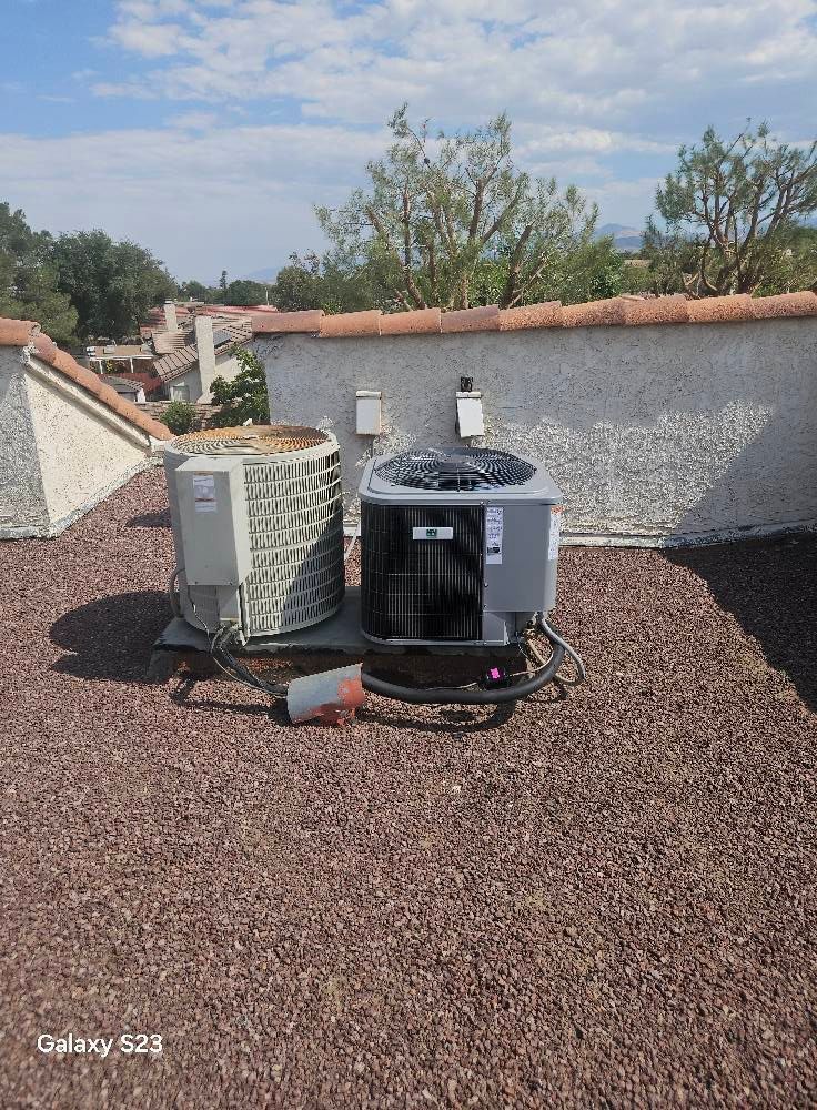 Two air conditioning units sit on a flat, gravel-covered roof against a backdrop of houses and trees.