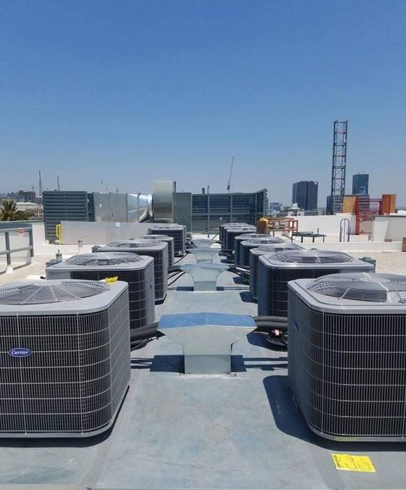 Multiple grey HVAC condenser units arranged in rows on a flat, sunlit building rooftop under a clear blue sky.