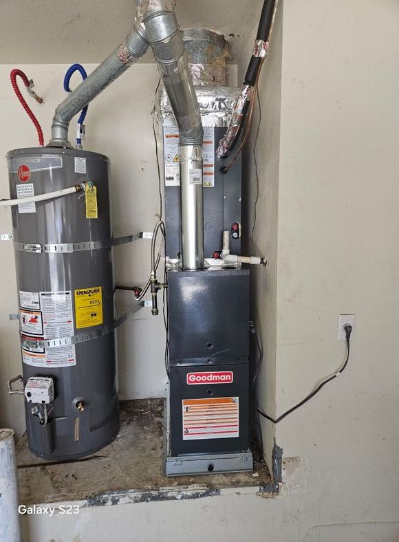 A gray water heater stands next to a tall, black Goodman furnace in a residential utility alcove.