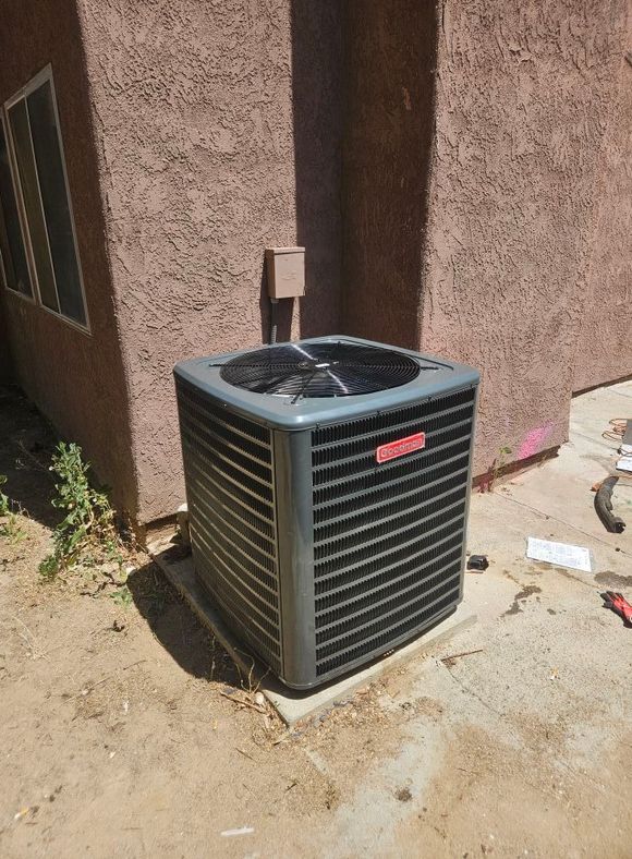 A gray Goodman air conditioning unit sits outside against a beige stucco wall.