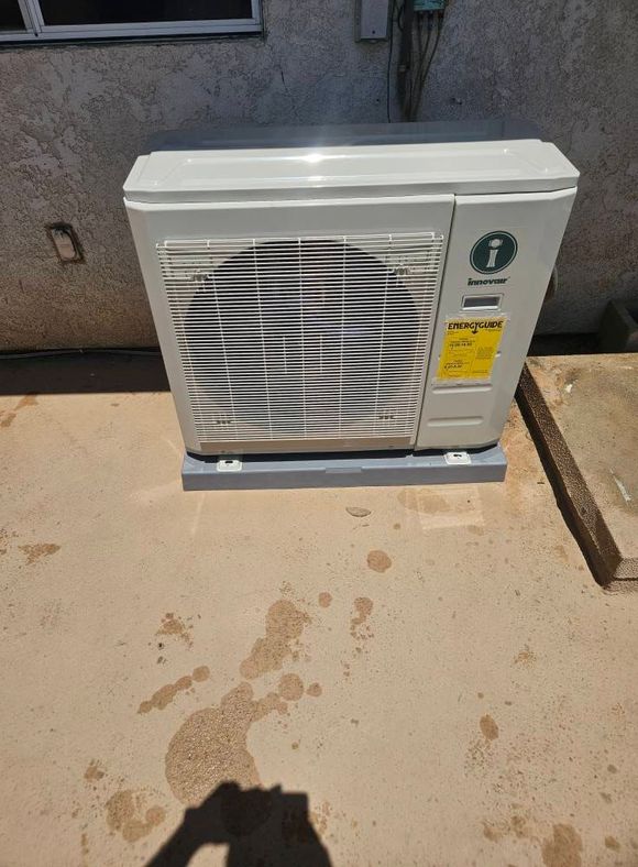 A white outdoor mini-split air conditioner unit sits on a concrete pad against a textured wall.