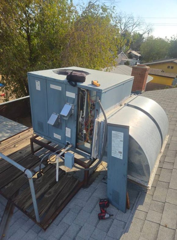 A grey rooftop HVAC unit with an open electrical panel and detached cover sitting on a wooden frame on a shingled roof.