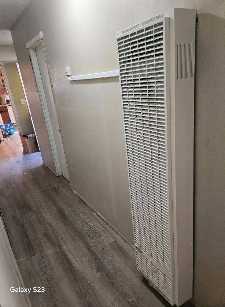A wall-mounted white heater in a hallway with grey wood-style flooring.
