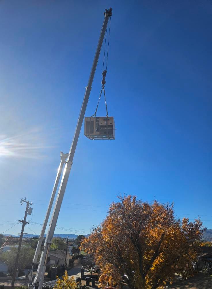 A crane suspends a large HVAC unit in the air against a clear blue sky, next to a tree with autumn foliage.