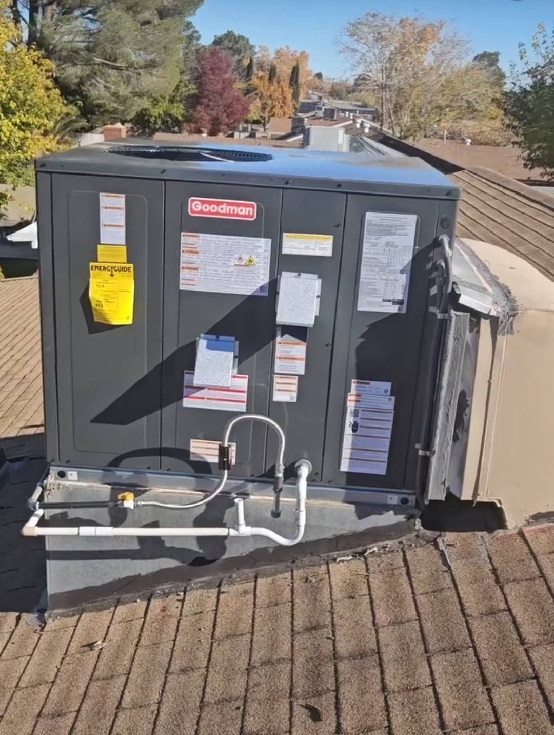 A gray Goodman HVAC unit sits on a residential shingled roof under a clear blue sky.