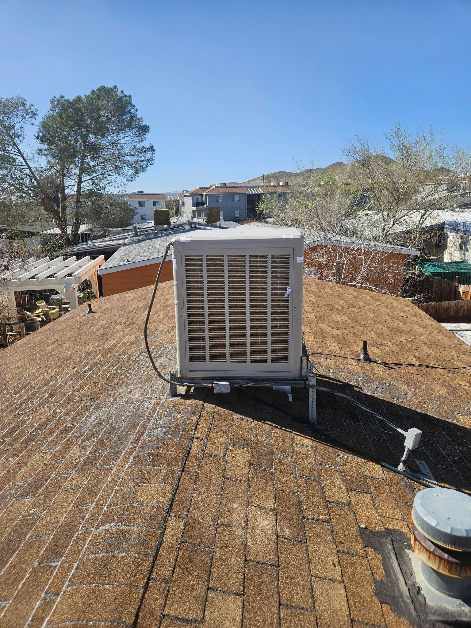 A residential swamp cooler sits on a shingled roof under a clear blue sky, with a suburban neighborhood in the background.