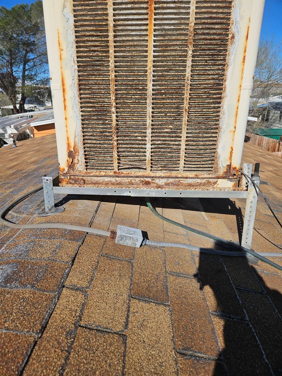 An old, rusted swamp cooler sitting on a shingled roof, connected by an electrical conduit and wire box.
