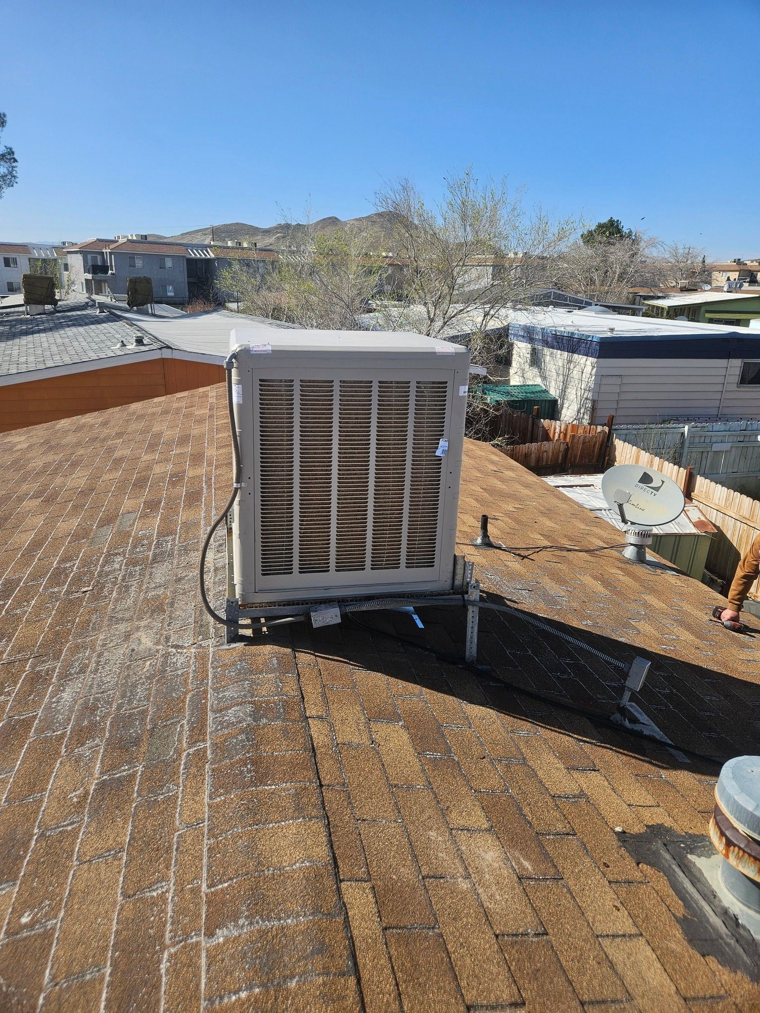 An evaporative cooler sits on a shingled residential roof under a bright blue sky.