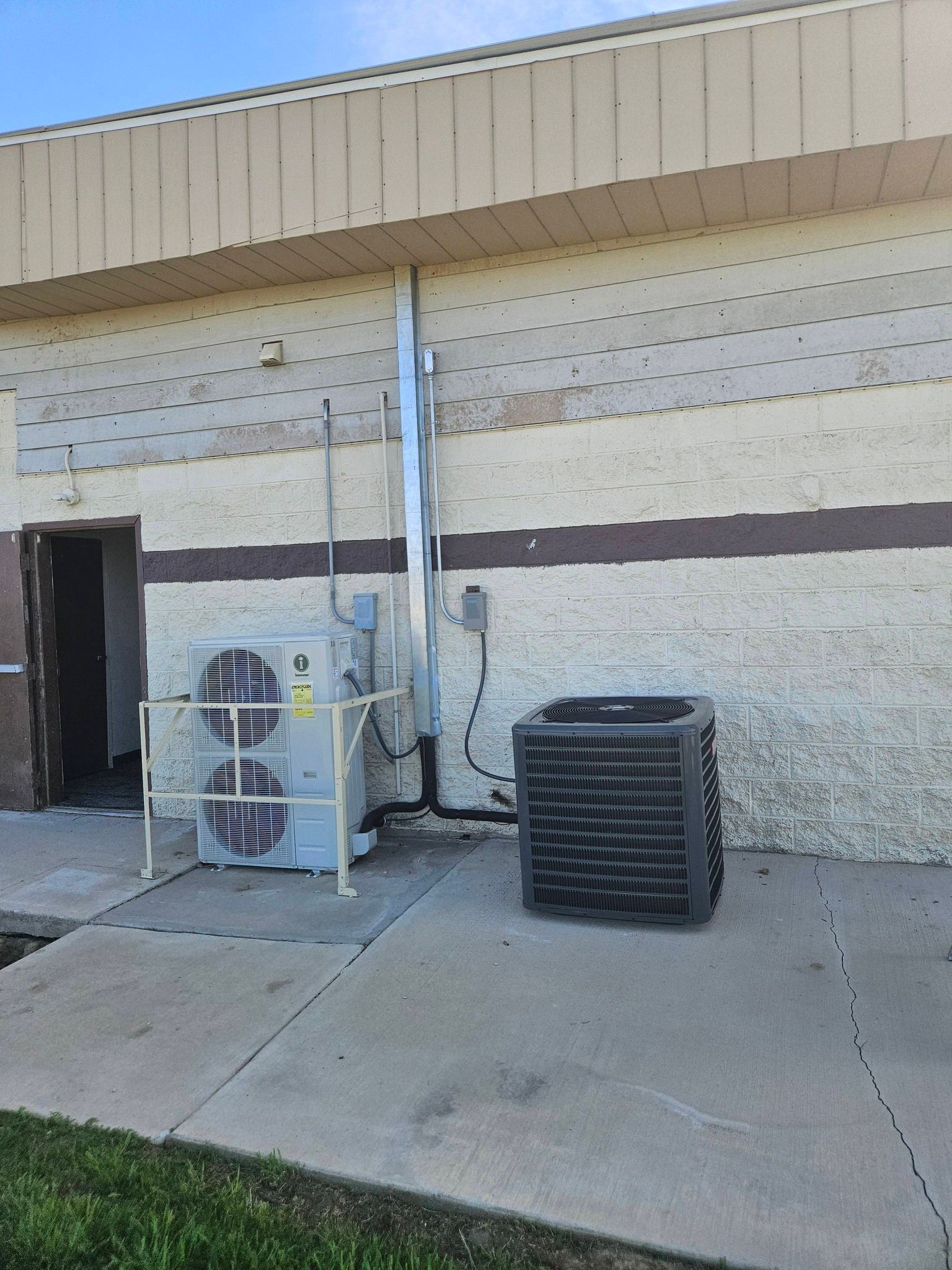 Two industrial HVAC units sit on a concrete pad against a beige, multi-textured wall near an open door.