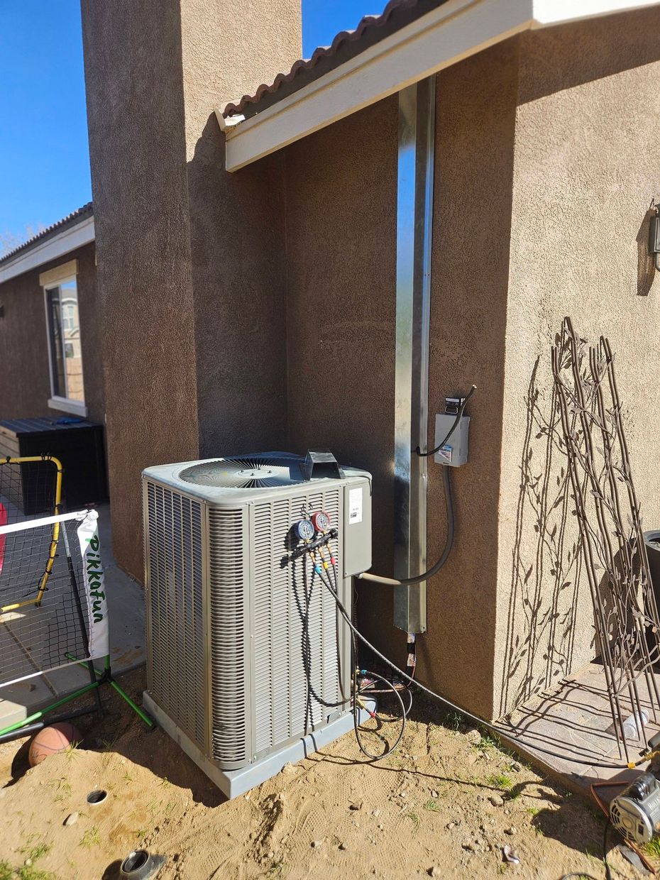 An air conditioning condenser unit sits on the sandy ground next to an exterior stucco wall with metal utility housing.