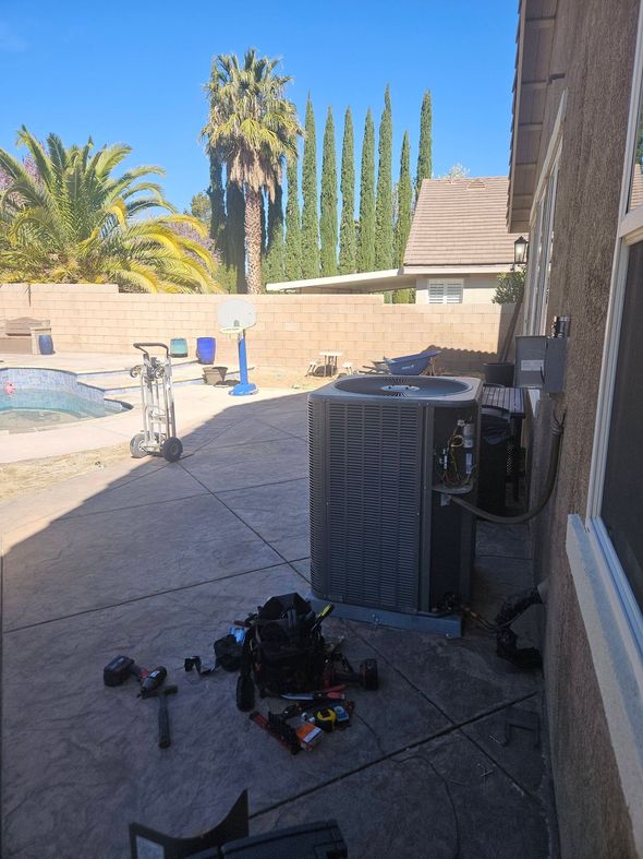 An HVAC condenser unit sits on a backyard patio next to tools and a bag, with a swimming pool in the background.