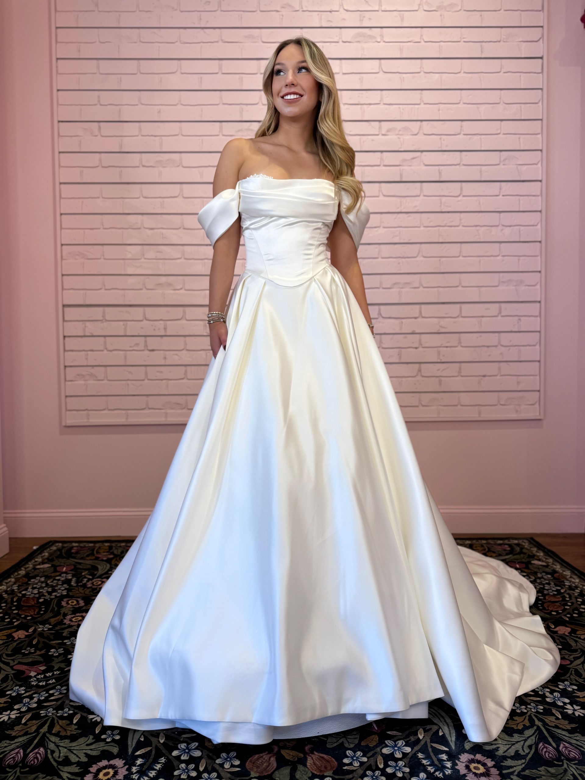 Woman in an off-the-shoulder white wedding gown smiles, posing against a pink brick wall.
