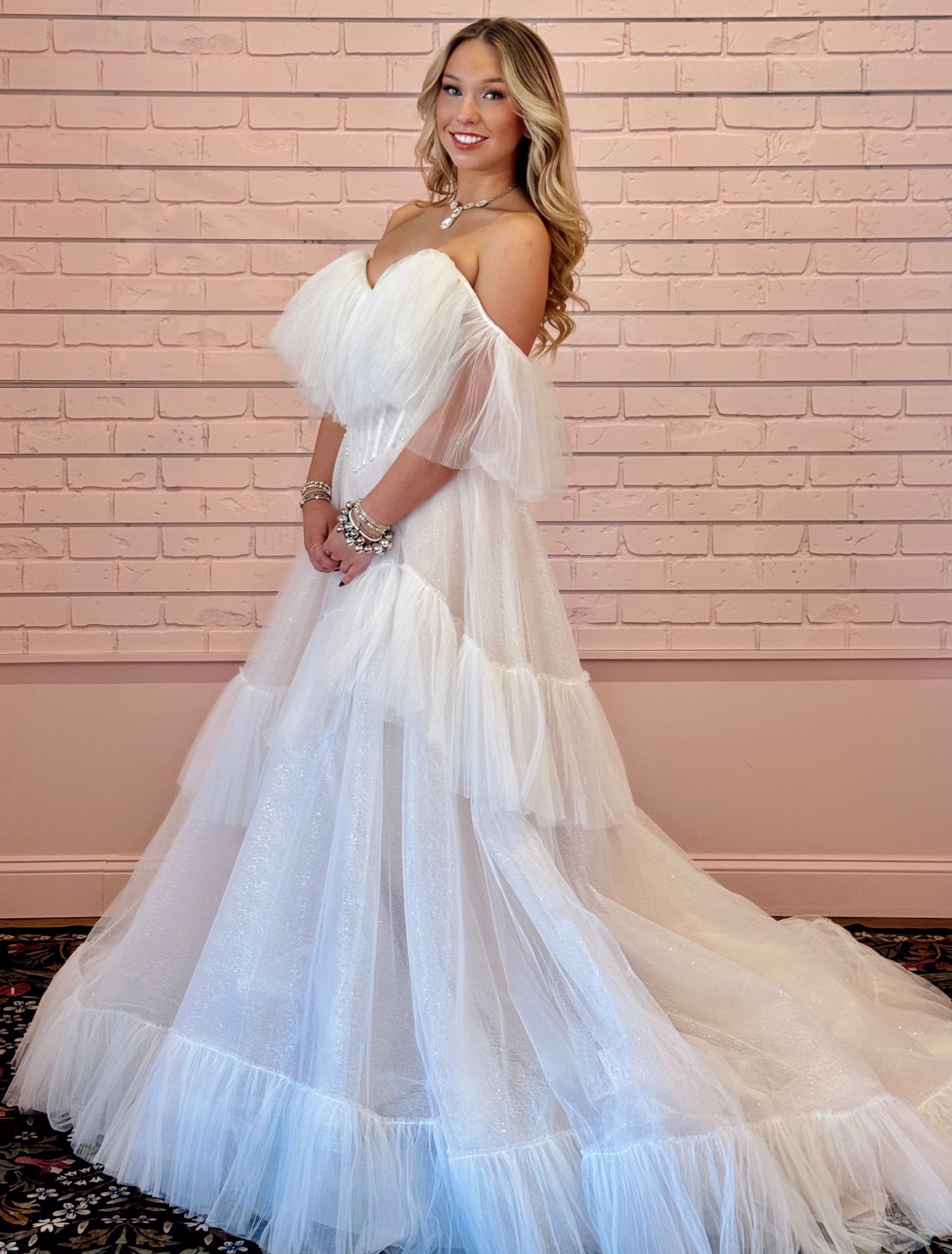 Woman in white off-the-shoulder gown stands smiling against a pink brick wall.