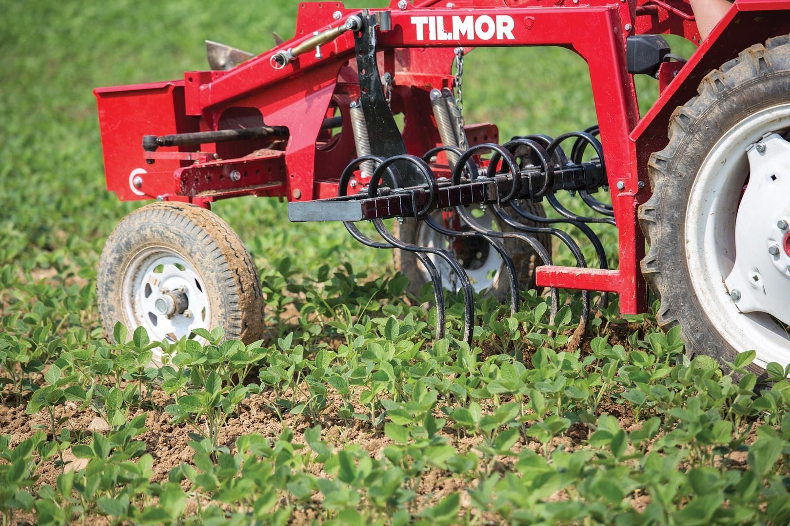Red Tilmor farm implement working in a green field, with a tractor wheel visible on the right