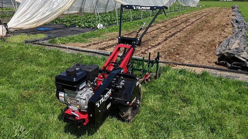 Red walk-behind tiller on grass beside a covered garden plot and dirt rows.