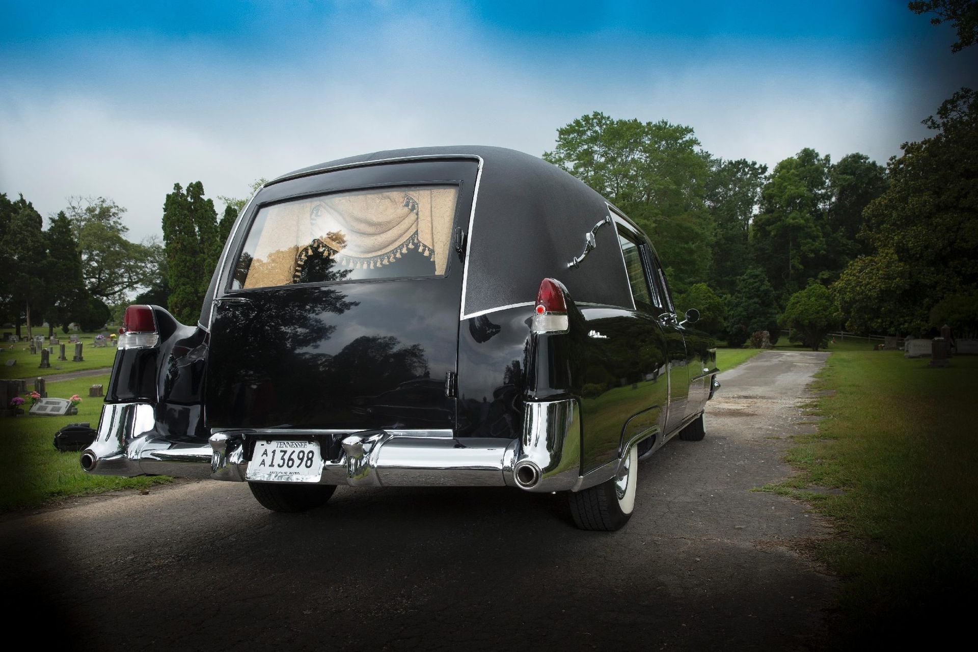 Black vintage hearse parked on a tree-lined road with its rear facing the viewer.