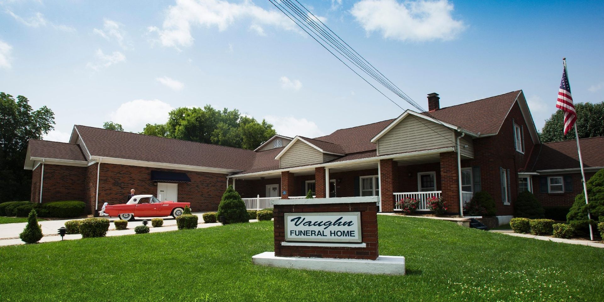 Brick assisted-living building with a sign, parked red car, and American flag in front