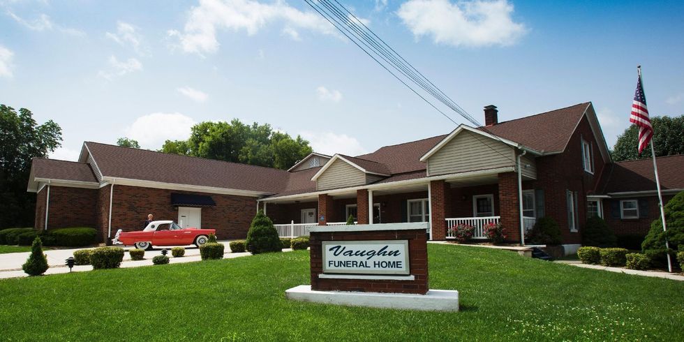 Brick assisted-living building with a sign, parked red car, and American flag in front