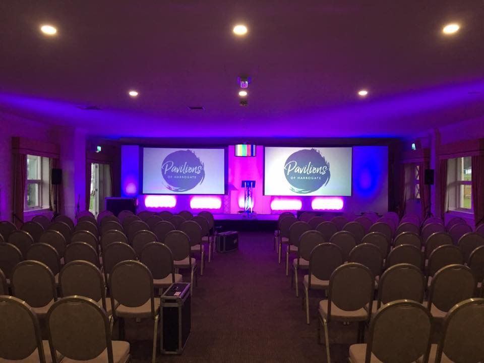 Two large screens and uplighting  at the front of a meeting room.Production services and equipment  for this corporate event was supplied by Exclusive Entertainments Harrogate,Yorkshire