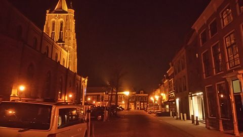 Een straat 's nachts met gloeiende oranje lampen, een verlichte kerktoren en geparkeerde auto's langs oude gebouwen.
